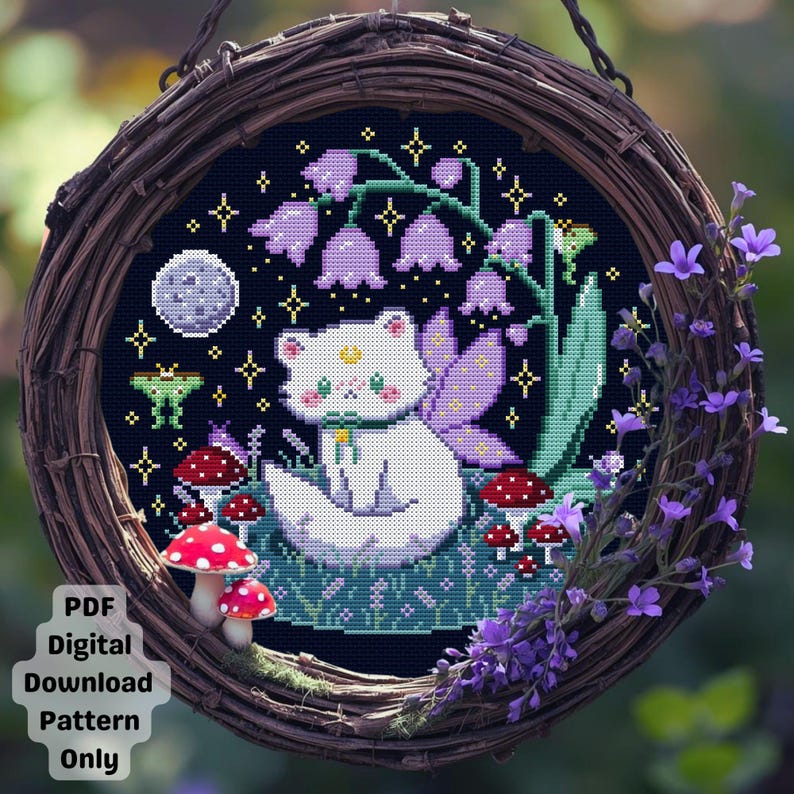 Op de afbeelding: Een kruissteekontwerp met een witte kat met vleugels in een fantasietuin. De sc&egrave;ne bevat paarse bloemen, rode paddenstoelen, een maan en sterren op een donkerblauwe achtergrond. Het ontwerp is omlijst in een geweven krans.
