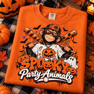 May include: An orange crewneck sweatshirt featuring a graphic of a monkey wearing a baseball cap and sunglasses, holding two baseball bats. The design includes pumpkins, bats, a ghost, and the text "SPOOKY Party Animals" in orange and white.