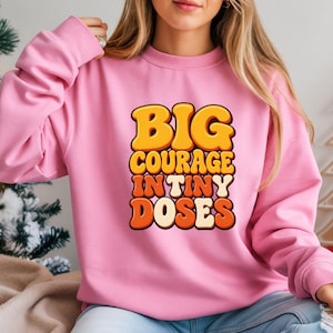 May include: A light pink long-sleeve sweatshirt featuring the text BIG COURAGE IN TINY DOSES printed in a stacked, bubbly, retro-style font. The lettering uses a gradient of yellow, orange, and cream colors with a dark outline.