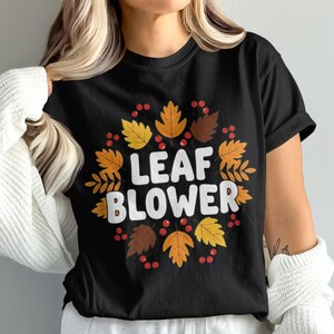 Peut inclure: T-shirt noir avec un motif sur le thème de l'automne. Le motif présente une couronne de feuilles d'automne jaunes, oranges et marron, entourant les mots "LEAF BLOWER" en blanc. Des baies rouges sont également incluses.