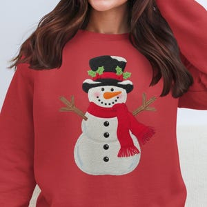 Peut inclure: Sweat-shirt rouge avec un motif de bonhomme de neige brodé. Le bonhomme de neige porte un chapeau haut de forme noir, une écharpe rouge et des bras en brindilles. Un vêtement d'hiver festif.