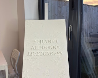 May include: A white canvas with textured surface, mounted on a wooden easel. The canvas features the engraved text: "YOU AND I ARE GONNA LIVE FOREVER". The easel is positioned in front of a glass door, with a glimpse of the outside.