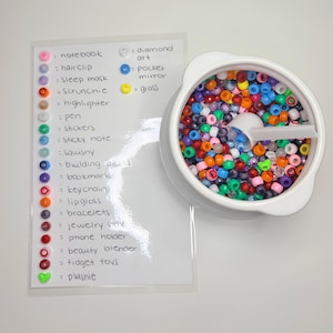 May include: A white container filled with colourful beads, next to a list of items with corresponding colour-coded dots. The list includes items like notebooks, hair clips, and keychains. The beads are in various colours.