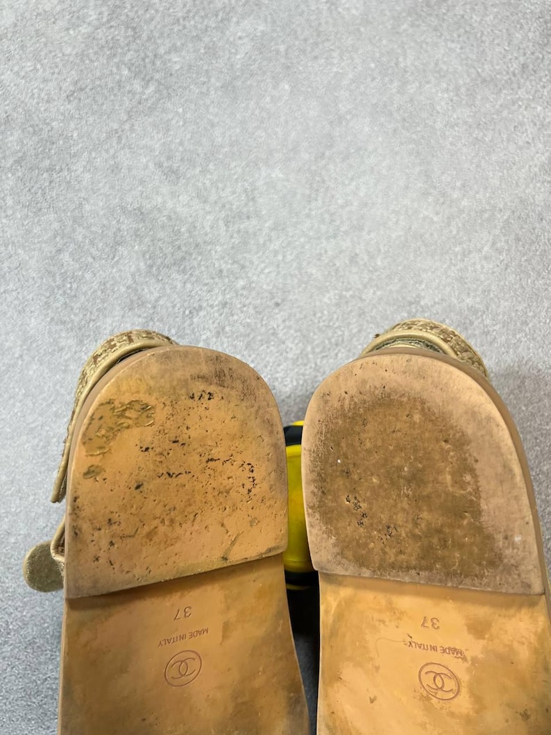 May include: A pair of beige sandals with a textured strap and a buckle closure. The soles are worn and show the size 37, and the brand logo. The sandals are made in Italy.
