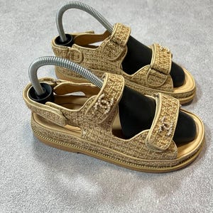 May include: A pair of beige sandals with adjustable straps and a woven texture. The sandals have a platform sole and feature decorative embellishments on the straps. Shoe trees are inserted in the sandals.