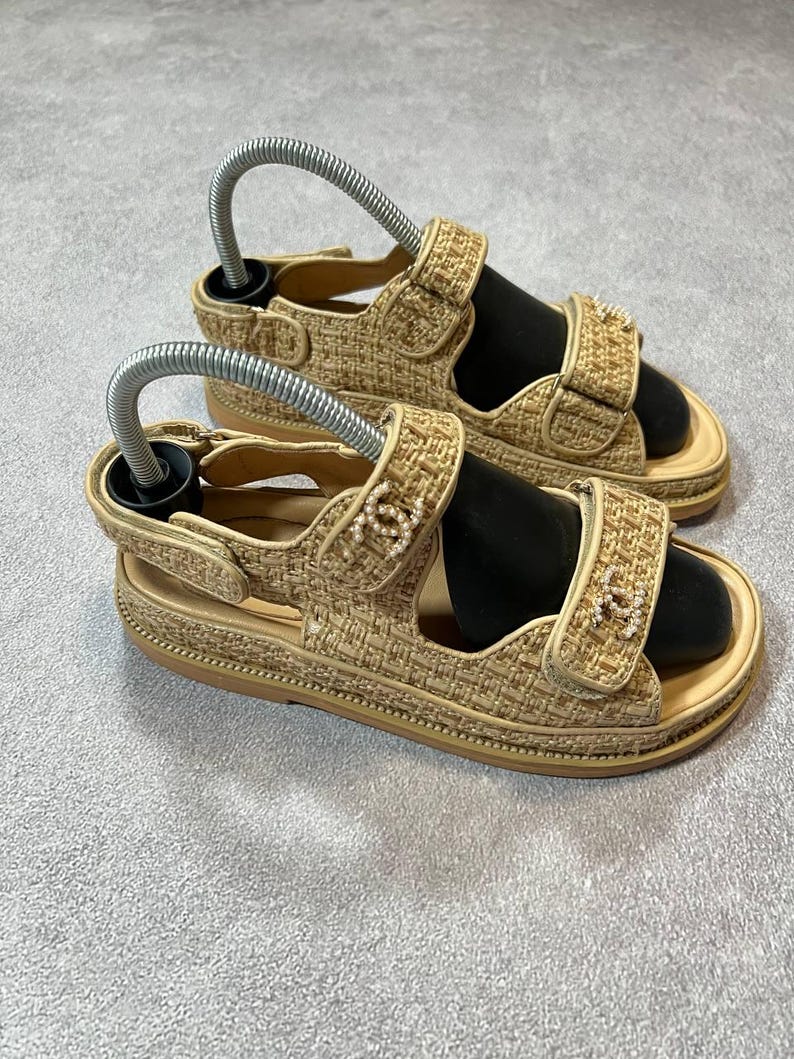 May include: A pair of beige sandals with adjustable straps and a woven texture. The sandals have a platform sole and feature decorative embellishments on the straps. Shoe trees are inserted in the sandals.