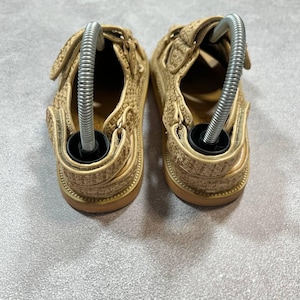 May include: A pair of beige sandals with hook-and-loop closures. The sandals have a woven texture and a brown sole. Metal shoe trees are inserted into each sandal.