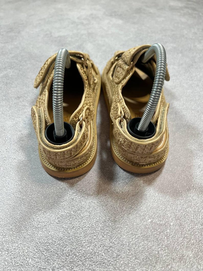 May include: A pair of beige sandals with hook-and-loop closures. The sandals have a woven texture and a brown sole. Metal shoe trees are inserted into each sandal.