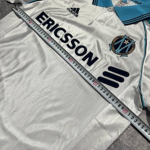Peut inclure: Maillot de football blanc avec des d&eacute;tails bleus, arborant le logo "ERICSSON" et la marque Adidas. Le maillot a un col et des bordures bleus, avec un &eacute;cusson d'&eacute;quipe sur la poitrine. Mesure du maillot avec un m&egrave;tre ruban.