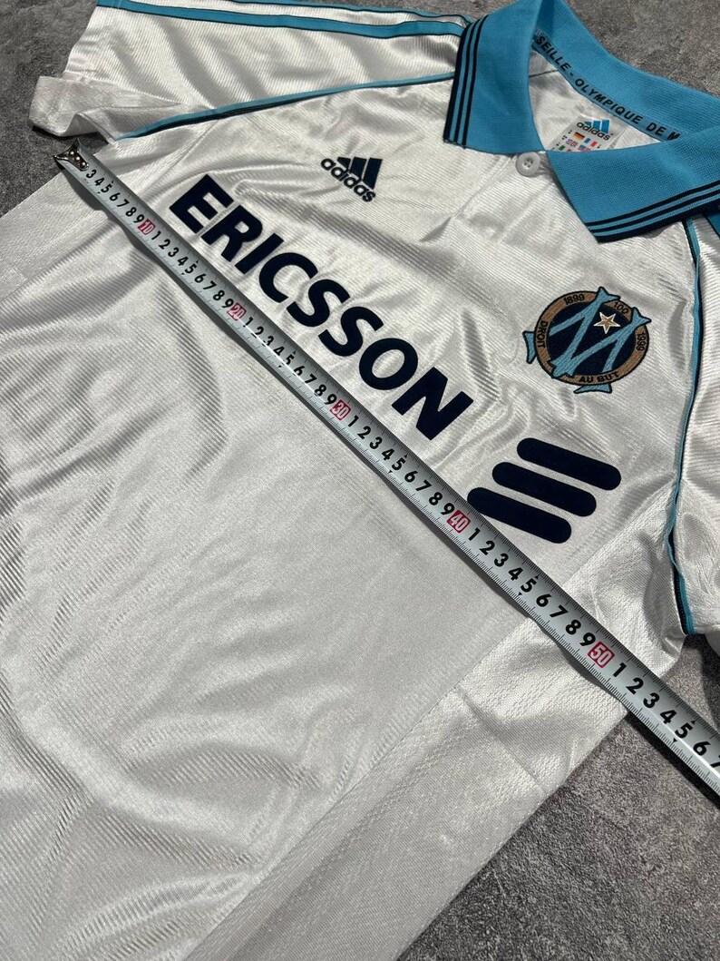 Peut inclure: Maillot de football blanc avec des d&eacute;tails bleus, arborant le logo "ERICSSON" et la marque Adidas. Le maillot a un col et des bordures bleus, avec un &eacute;cusson d'&eacute;quipe sur la poitrine. Mesure du maillot avec un m&egrave;tre ruban.