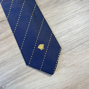 Puede incluir: Corbata azul marino con rayas verticales punteadas amarillas. Un emblema dorado está cerca de la parte inferior. La corbata está hecha de una tela acanalada y se muestra sobre una superficie de madera clara.