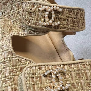 May include: Close-up of beige woven sandals with gold-tone hardware. The sandals feature a woven texture and a gold-tone logo embellished with small pearls on the straps. The interior is a light beige color.