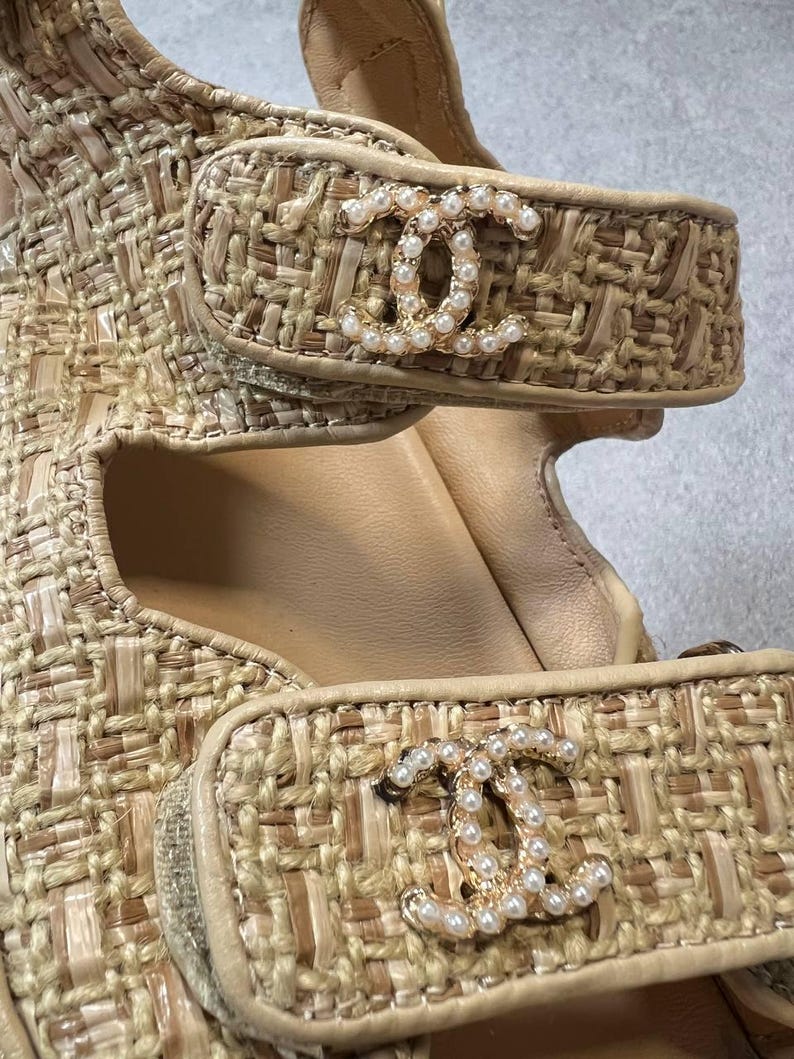 May include: Close-up of beige woven sandals with gold-tone hardware. The sandals feature a woven texture and a gold-tone logo embellished with small pearls on the straps. The interior is a light beige color.
