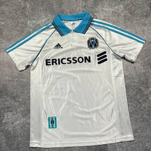 Peut inclure: Maillot de football blanc avec des d&eacute;tails bleu clair, le logo Adidas et le mot "ERICSSON" en noir. Col bleu et &eacute;cusson de l'Olympique de Marseille. Tissu l&eacute;ger.
