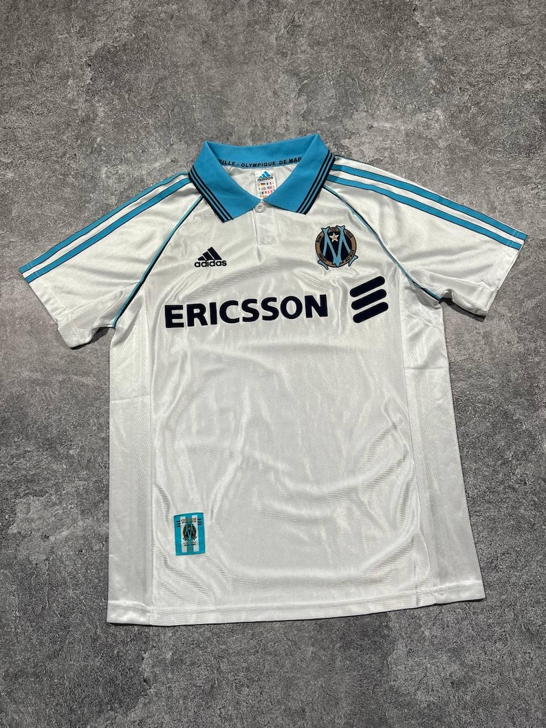 Peut inclure: Maillot de football blanc avec des d&eacute;tails bleu clair, le logo Adidas et le mot "ERICSSON" en noir. Col bleu et &eacute;cusson de l'Olympique de Marseille. Tissu l&eacute;ger.