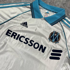 Peut inclure: Maillot de football blanc avec des d&eacute;tails bleu clair, notamment le logo Adidas et le mot "ERICSSON" en bleu marine. Le maillot a un col et des rayures bleu clair, ainsi qu'un &eacute;cusson d'&eacute;quipe.