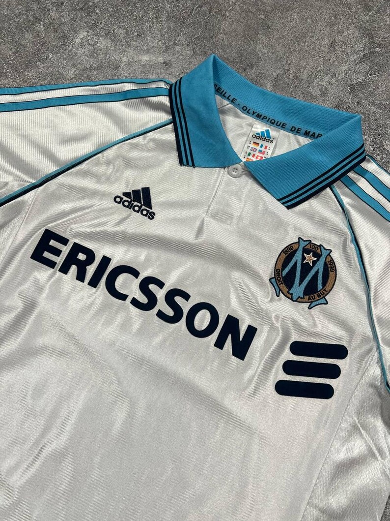 Peut inclure: Maillot de football blanc avec des d&eacute;tails bleu clair, notamment le logo Adidas et le mot "ERICSSON" en bleu marine. Le maillot a un col et des rayures bleu clair, ainsi qu'un &eacute;cusson d'&eacute;quipe.