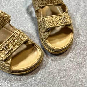 May include: Beige woven sandals with adjustable straps. The sandals feature a gold logo on the straps and a beige sole. The sandals are designed with a textured woven material.