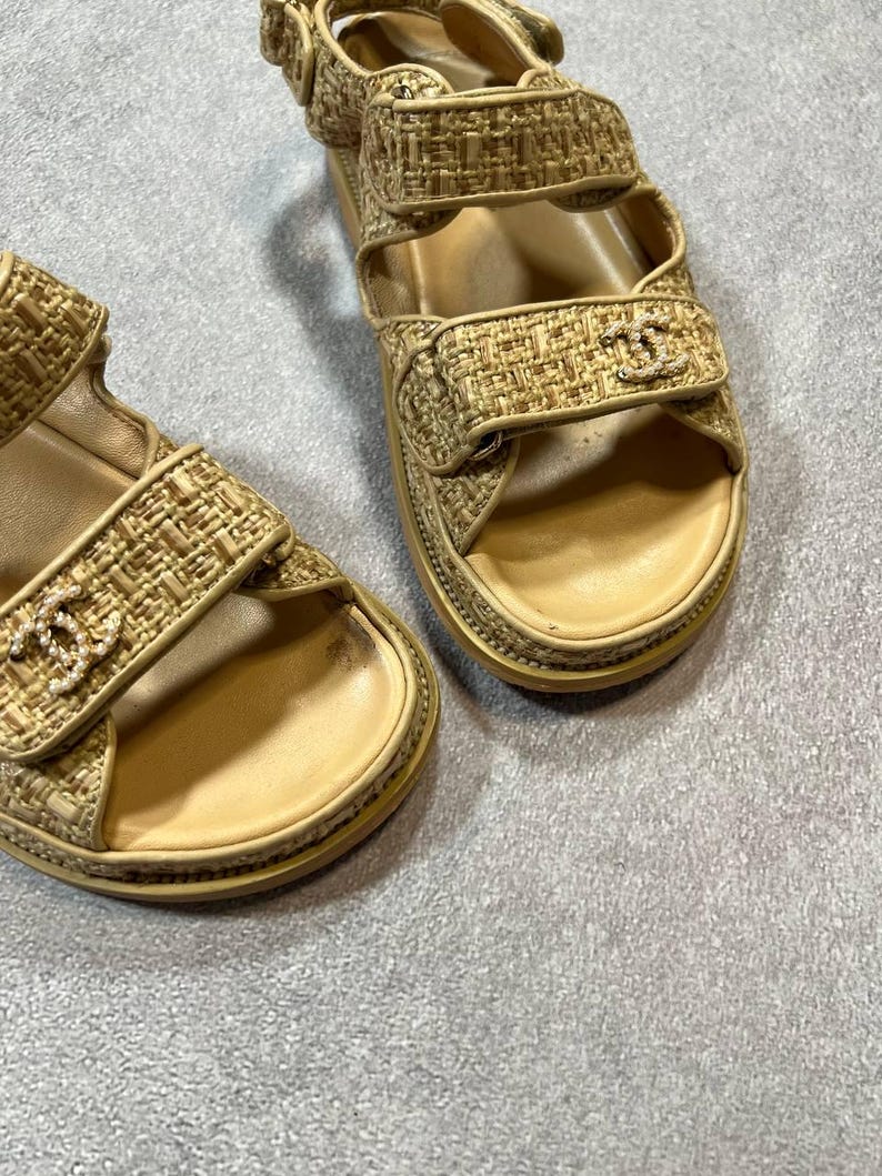 May include: Beige woven sandals with adjustable straps. The sandals feature a gold logo on the straps and a beige sole. The sandals are designed with a textured woven material.