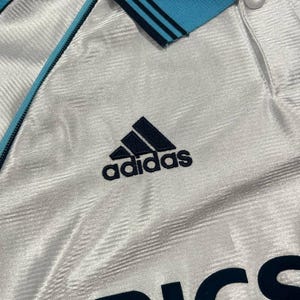Peut inclure: Maillot de sport blanc avec col et bordures bleu clair. Comporte le logo Adidas bleu marine et le mot "ERICA" en lettres majuscules bleu marine. Le maillot pr&eacute;sente un motif textur&eacute;.