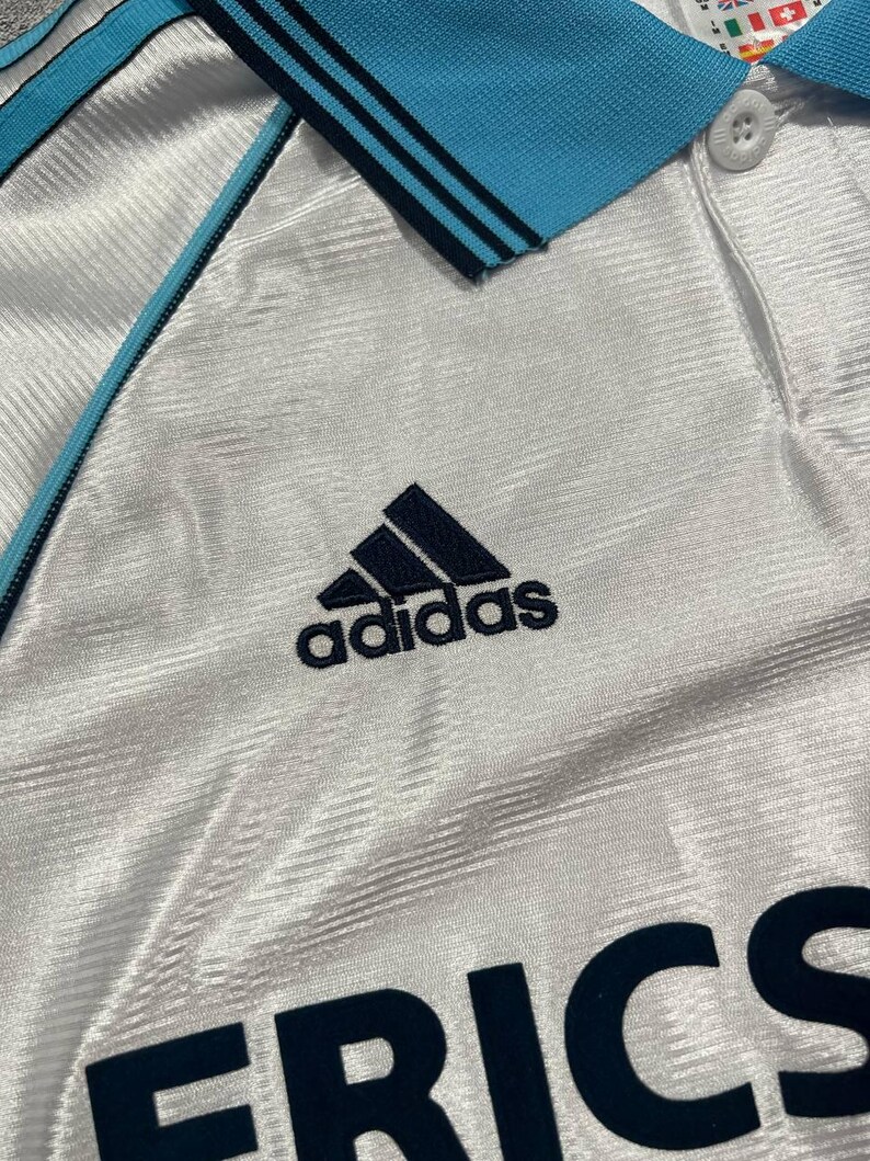 Peut inclure: Maillot de sport blanc avec col et bordures bleu clair. Comporte le logo Adidas bleu marine et le mot "ERICA" en lettres majuscules bleu marine. Le maillot pr&eacute;sente un motif textur&eacute;.