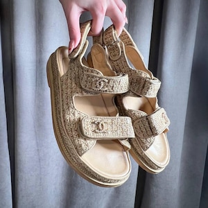 May include: Beige and tan sandals with adjustable straps. The sandals have a woven texture and a platform sole. The straps feature a logo detail. The sandals are held up by a hand.