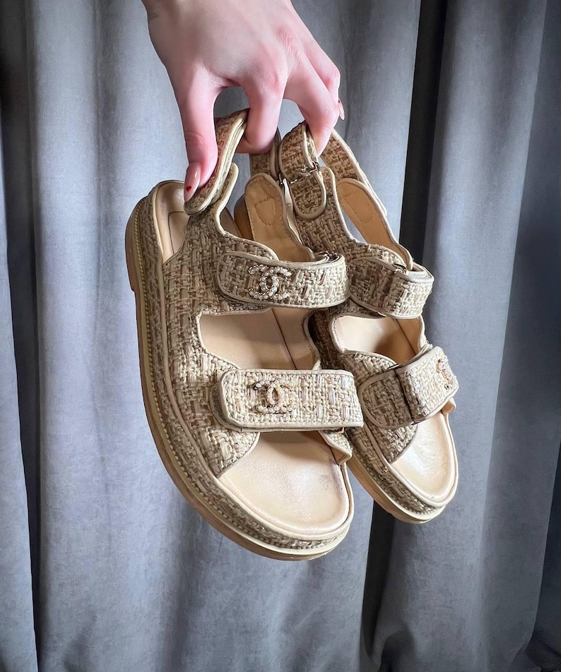 May include: Beige and tan sandals with adjustable straps. The sandals have a woven texture and a platform sole. The straps feature a logo detail. The sandals are held up by a hand.