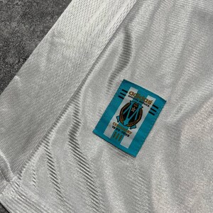 Peut inclure: Gros plan sur un maillot blanc avec un motif textur&eacute;. Une &eacute;tiquette rectangulaire bleue avec le logo Adidas et le texte "OFFICIAL REPLICA" est cousue sur le maillot. L'&eacute;tiquette comporte &eacute;galement un &eacute;cusson et le texte "CENTENARY 1899-1999".