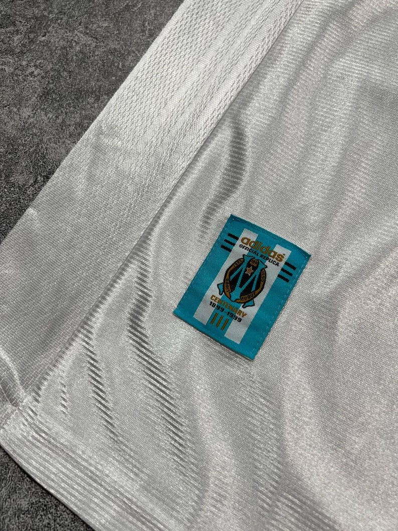 Peut inclure: Gros plan sur un maillot blanc avec un motif textur&eacute;. Une &eacute;tiquette rectangulaire bleue avec le logo Adidas et le texte "OFFICIAL REPLICA" est cousue sur le maillot. L'&eacute;tiquette comporte &eacute;galement un &eacute;cusson et le texte "CENTENARY 1899-1999".