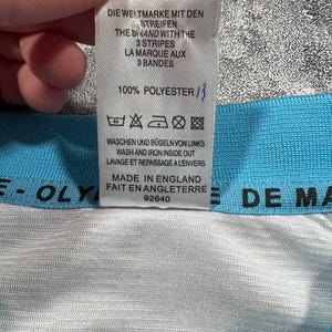 Peut inclure: Gros plan sur une &eacute;tiquette blanche avec du texte noir, indiquant les instructions de lavage et la mention "Made in England". L'&eacute;tiquette est attach&eacute;e &agrave; un tissu bleu clair et blanc. Le texte comprend "100% Polyester."
