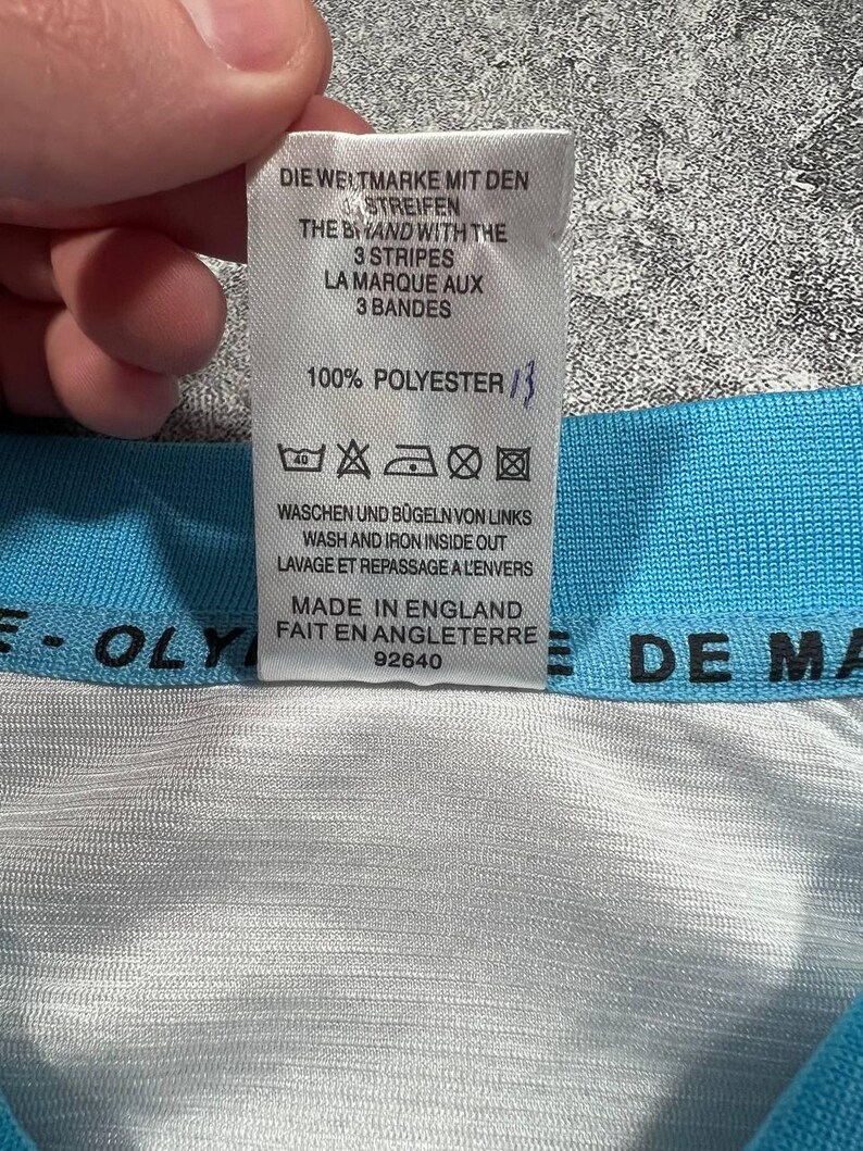 Peut inclure: Gros plan sur une &eacute;tiquette blanche avec du texte noir, indiquant les instructions de lavage et la mention "Made in England". L'&eacute;tiquette est attach&eacute;e &agrave; un tissu bleu clair et blanc. Le texte comprend "100% Polyester."
