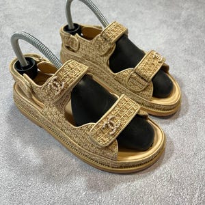 May include: A pair of beige fabric sandals with adjustable straps. The sandals have a woven texture and a small logo on the strap. The soles are a light beige color. The sandals are displayed on a gray surface.
