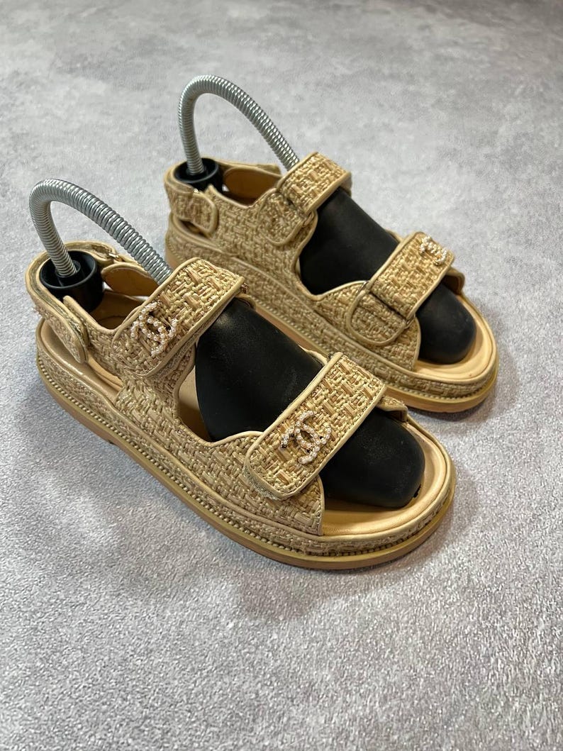 May include: A pair of beige fabric sandals with adjustable straps. The sandals have a woven texture and a small logo on the strap. The soles are a light beige color. The sandals are displayed on a gray surface.