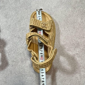 May include: Tan sandal with adjustable straps. The sandal has a woven texture and a logo detail. The sandal measures approximately 9 inches in length.