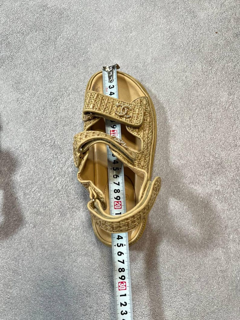 May include: Tan sandal with adjustable straps. The sandal has a woven texture and a logo detail. The sandal measures approximately 9 inches in length.