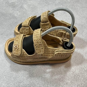 May include: Beige woven sandals with adjustable straps. The sandals have a textured design and a light brown sole. The straps feature a logo detail. The sandals are displayed on a gray surface.