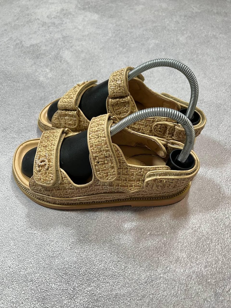 May include: Beige woven sandals with adjustable straps. The sandals have a textured design and a light brown sole. The straps feature a logo detail. The sandals are displayed on a gray surface.