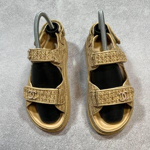 May include: A pair of beige sandals with a woven texture. The sandals have adjustable straps and a small logo on the front strap. The soles are a light beige color. The sandals are displayed on a gray surface.