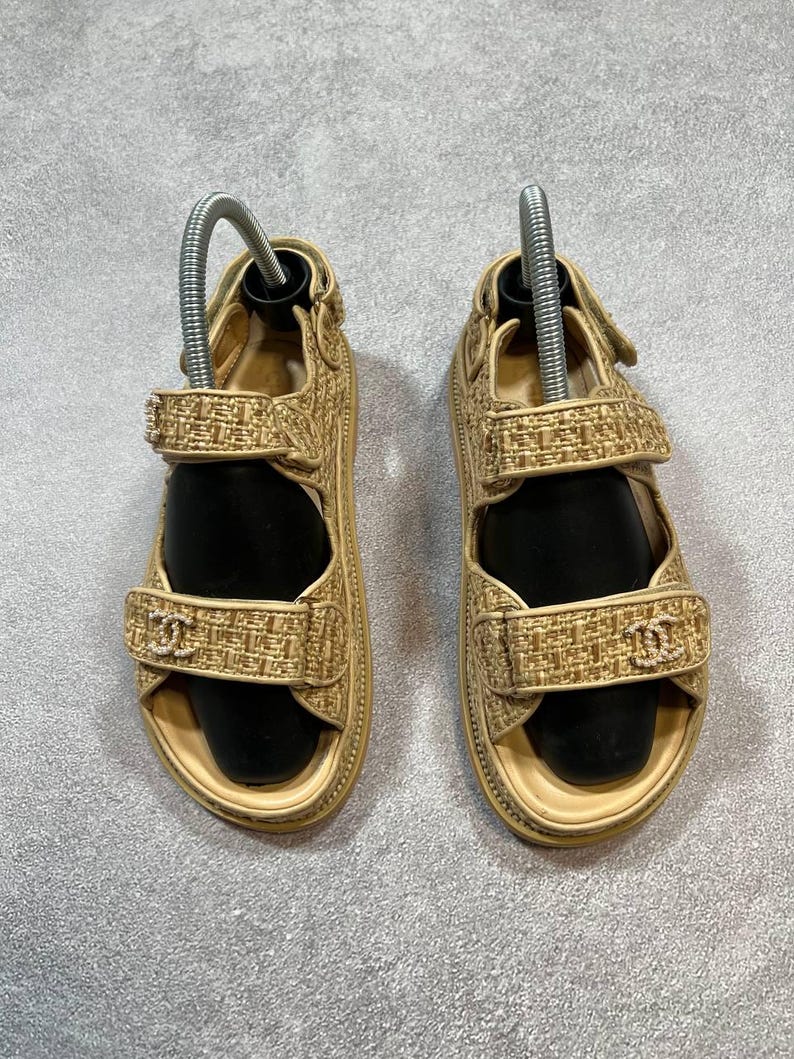 May include: A pair of beige sandals with a woven texture. The sandals have adjustable straps and a small logo on the front strap. The soles are a light beige color. The sandals are displayed on a gray surface.