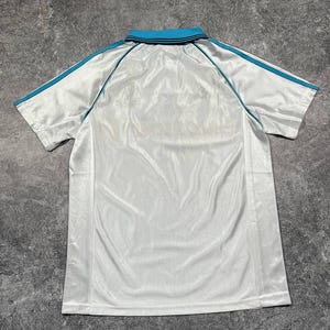 Peut inclure: Polo blanc avec col turquoise et rayures turquoise sur les manches. Le t-shirt est fait d'une mati&egrave;re brillante. Le t-shirt est pos&eacute; &agrave; plat sur une surface grise.