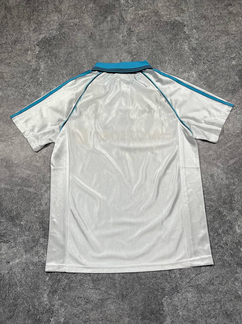 Peut inclure: Polo blanc avec col turquoise et rayures turquoise sur les manches. Le t-shirt est fait d'une mati&egrave;re brillante. Le t-shirt est pos&eacute; &agrave; plat sur une surface grise.