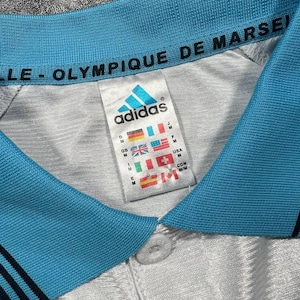 Peut inclure: Gros plan sur un maillot de football Adidas blanc et bleu clair. Le maillot a un col bleu clair avec le texte "OLYMPIQUE DE MARSEILLE". Le logo Adidas est visible sur l'&eacute;tiquette, ainsi que divers drapeaux de pays.