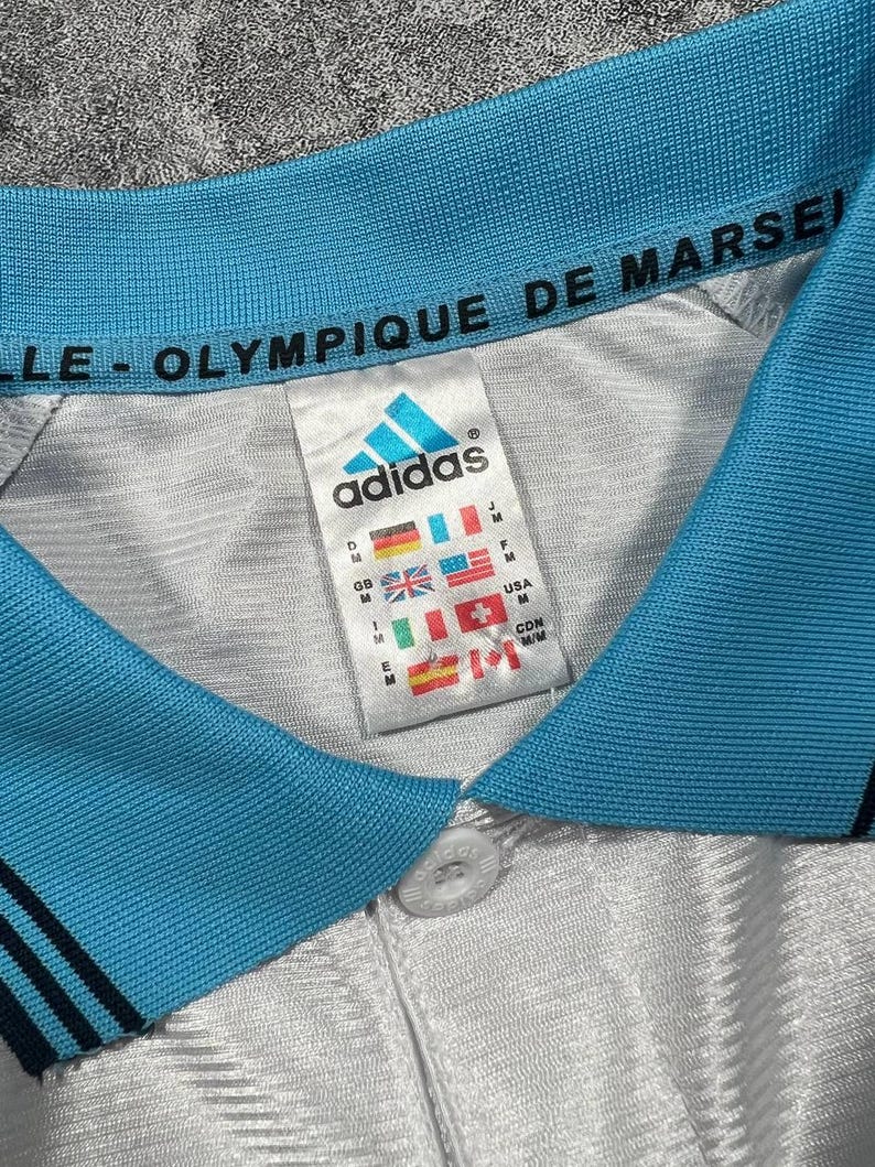 Peut inclure: Gros plan sur un maillot de football Adidas blanc et bleu clair. Le maillot a un col bleu clair avec le texte "OLYMPIQUE DE MARSEILLE". Le logo Adidas est visible sur l'&eacute;tiquette, ainsi que divers drapeaux de pays.
