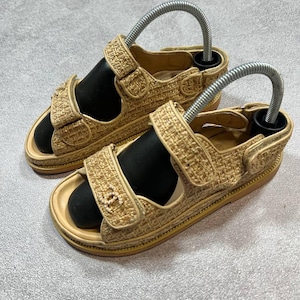 May include: A pair of beige woven sandals with adjustable straps and a black footbed. The sandals have a gold-colored trim and a logo on the strap. The sandals are displayed with metal shoe trees.