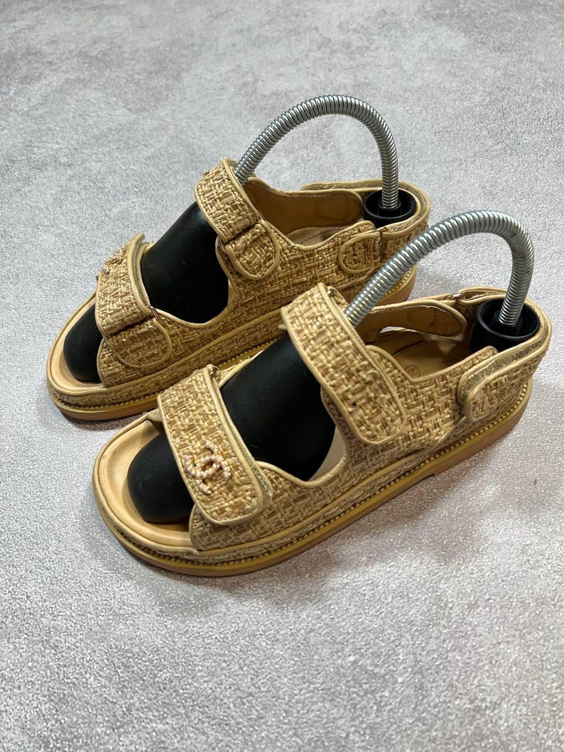 May include: A pair of beige woven sandals with adjustable straps and a black footbed. The sandals have a gold-colored trim and a logo on the strap. The sandals are displayed with metal shoe trees.