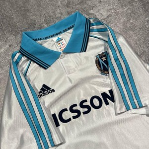 Peut inclure: Maillot de football blanc avec des d&eacute;tails bleu clair, avec le logo Adidas et le mot "ICSSON". Le maillot a un col bleu clair avec des rayures noires et le texte "VILLE-OLYMPIQUE DE MAR".