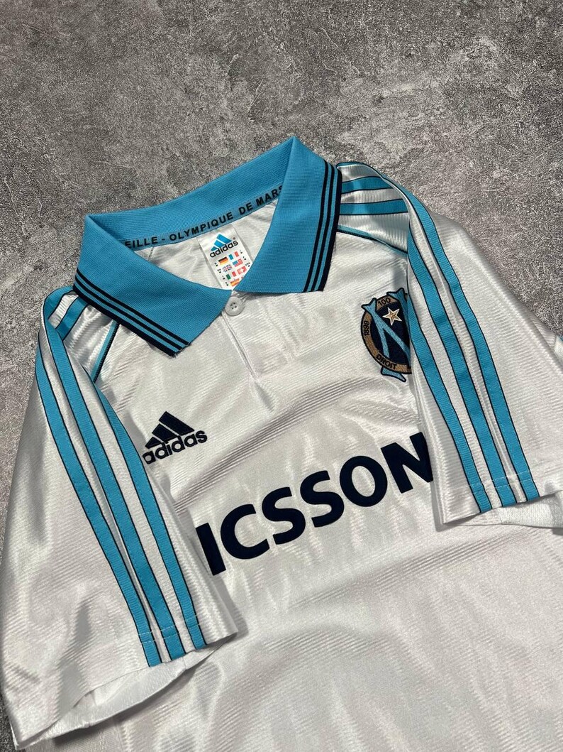 Peut inclure: Maillot de football blanc avec des d&eacute;tails bleu clair, avec le logo Adidas et le mot "ICSSON". Le maillot a un col bleu clair avec des rayures noires et le texte "VILLE-OLYMPIQUE DE MAR".