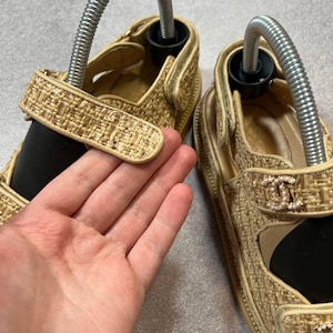 May include: Tan woven sandals with a gold trim and a decorative logo on the strap. The sandals have a velcro strap closure and are shown with shoe trees inserted. The sandals are on a neutral background.