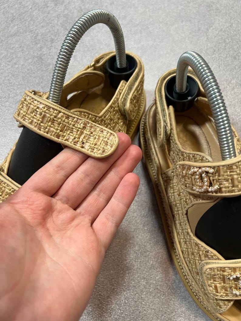 May include: Tan woven sandals with a gold trim and a decorative logo on the strap. The sandals have a velcro strap closure and are shown with shoe trees inserted. The sandals are on a neutral background.