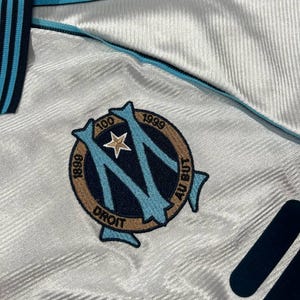 Peut inclure: Gros plan sur un maillot de football blanc et bleu clair. Le maillot pr&eacute;sente un logo d'&eacute;quipe avec le texte "1899", "1999" et "Droit". Le maillot a des accents bleu clair et bleu marine.